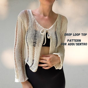 Op de afbeelding: Een wit gehaakt topje met een drop loop design. Het topje heeft een strik bij de halslijn en lange mouwen. De tekst "DROP LOOP TOP PATTERN FOR ADDI/SENTRO" is op de afbeelding gedrukt.