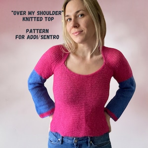Puede incluir: Un top de punto rosa con mangas azules. El top tiene un escote redondo y es corto. El texto "Over My Shoulder Knitted Top Pattern for Addi/Sentro" está escrito encima de la imagen.