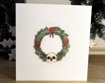 May include: A white card with a green wreath decorated with red bows and a white skull in the center. The wreath is on a white background.