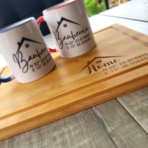 Könnte beinhalten: Zwei weiße Keramikbecher mit blauem und rotem Rand. Jeder Becher hat ein Hausdesign mit dem Text "Bauherrin" und Koordinaten. Die Becher stehen auf einem Holzbrett mit dem Text "Home" und Koordinaten.