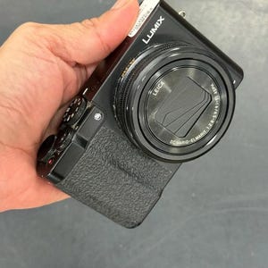 Könnte beinhalten: Eine schwarze Lumix Digitalkamera mit einem Leica DC Vario-Elmarit 1:2.8-5.9/9.1-91 ASPH Objektiv. Das Kameragehäuse verfügt über einen strukturierten Griff an der Vorderseite, ein Einstellrad oben und einen Auslöser mit rotem Punkt.