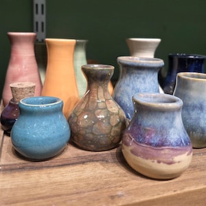 Może przedstawiać: Kolekcja małych, ręcznie robionych wazonów ceramicznych w różnych kolorach, w tym niebieskim, pomarańczowym i różowym. Niektóre wazony mają błyszczące wykończenie, inne wyglądają na teksturowane. Wazony są ułożone na drewnianej powierzchni.