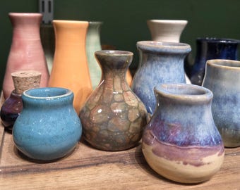 May include: A collection of small, handcrafted ceramic vases in various colors, including blue, orange, and pink. Some vases have a glossy finish, while others have a textured appearance. The vases are arranged on a wooden surface.