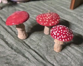 May include: Three ceramic mushroom figurines with red caps and white spots. The mushroom stems are beige. The mushrooms are arranged on a green fabric surface. The caps have a glossy finish.
