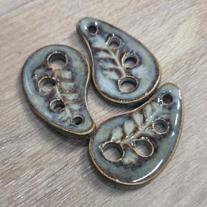 May include: Three teardrop-shaped ceramic buttons with a glossy, mottled blue-green glaze. Each button features a debossed fern leaf pattern and four circular holes arranged along the centre for attachment to fabric or other materials.
