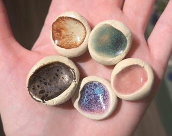 May include: A collection of five small, ceramic, pebble-shaped dishes. Each dish features a different interior color, including brown, green, black, blue, and pink, with a creamy white border. The dishes are held in a hand.