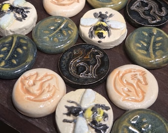 May include: A collection of small, round ceramic tiles with various embossed designs. The tiles feature images of bees, leaves, and stylized animals in shades of cream, black, green, and orange. The tiles are arranged on a wooden surface.