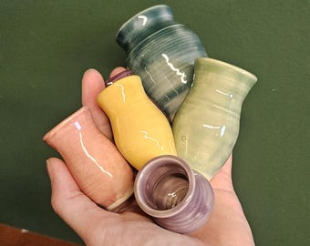 May include: A collection of small, handmade ceramic vases in various colors, including pink, yellow, green, purple, and teal. The vases have a smooth, glossy finish and a rounded shape, held in a hand against a green background.