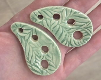 May include: Two teardrop-shaped ceramic buttons in a pale sage green glaze. Each button features a glossy finish with an embossed leaf pattern and three circular holes arranged in a line for attachment to fabric or other materials.
