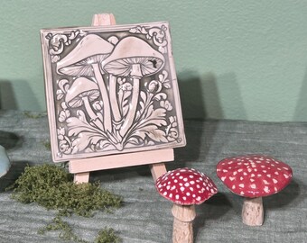 May include: A ceramic tile with a relief of three mushrooms on a small wooden easel. Two red and white spotted mushroom figurines are next to the tile. The background is a light green color.