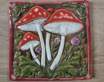 May include: A ceramic tile featuring a detailed illustration of three red mushrooms with white spots and white stems. The mushrooms are set against a brown background with green foliage and purple accents, framed by a red border.