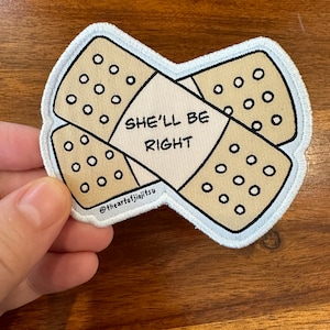 Puede incluir: Parche bordado con forma de venda con el texto "SHE'LL BE RIGHT" en negro. El parche es beige con contornos negros y costuras blancas. Tiene un borde blanco y el texto "@theartofjijitsu".