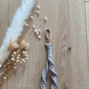 Op de afbeelding: Een lichtgrijze macram&eacute; sleutelhanger met een goudkleurige sluiting. De sleutelhanger is gemaakt van geweven katoenen koord in een spiraalvormig ontwerp. De achtergrond omvat gedroogde bloemen en een houten oppervlak.
