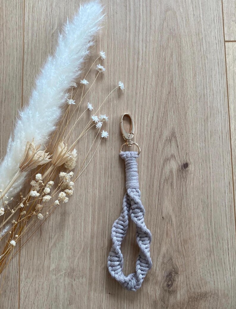 Op de afbeelding: Een lichtgrijze macram&eacute; sleutelhanger met een goudkleurige sluiting. De sleutelhanger is gemaakt van geweven katoenen koord in een spiraalvormig ontwerp. De achtergrond omvat gedroogde bloemen en een houten oppervlak.