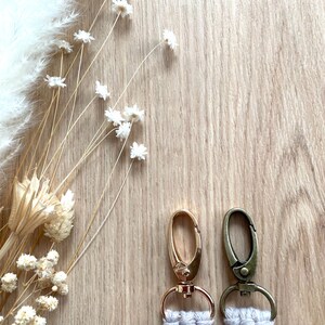 Op de afbeelding: Twee sleutelhangers met goud- en bronskleurige sluitingen en witte macram&eacute; details. De sleutelhangers staan tegen een lichte houtnerf achtergrond, met gedroogde bloemen en grassen in de linkerbovenhoek.