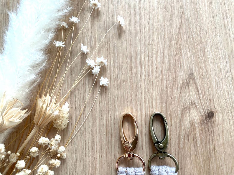 Op de afbeelding: Twee sleutelhangers met goud- en bronskleurige sluitingen en witte macram&eacute; details. De sleutelhangers staan tegen een lichte houtnerf achtergrond, met gedroogde bloemen en grassen in de linkerbovenhoek.
