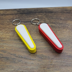 Könnte beinhalten: Zwei Schlüsselanhänger mit rechteckigem Design. Einer ist gelb mit weißer Mitte, der andere rot mit weißer Mitte. Jeder Schlüsselanhänger hat einen silbernen Ring.
