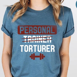 May include: A heather deep teal t-shirt with the text "PERSONAL TRAINER TORTURER" in white letters with a red box around the text. The word "TRAINER" is crossed out. A white dumbbell icon is below the text.