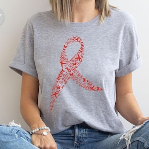 May include: Heather grey t-shirt with a red ribbon design. The ribbon is made up of words like "Hope", "Strength", "Love", and "Faith".