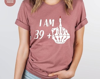 Puede incluir: Camiseta rosa polvorienta con mangas enrolladas. La camiseta presenta el texto "I AM 39+" sobre una mano esquelética blanca que muestra el dedo medio. En la esquina superior izquierda, un pequeño círculo con el texto "heather mavue".