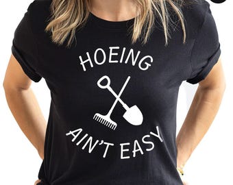 Puede incluir: Camiseta negra con la frase blanca "HOEING AIN'T EASY" y un diseño cruzado de rastrillo y pala. La camiseta tiene cuello redondo y parece estar hecha de un material suave. La palabra "negro" está en un círculo en la esquina superior derecha.