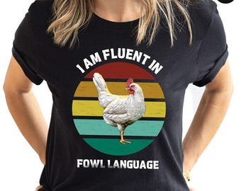 May include: Black t-shirt with a graphic of a white chicken inside a colorful circle. The text "I AM FLUENT IN FOWL LANGUAGE" is printed above and below the chicken illustration. The shirt is a crew neck.