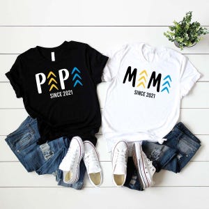 May include: Two black and white t-shirts with the words "Papa" and "Mama" in black with yellow and blue arrows. The text "Since 2021" is printed below the words on each shirt.