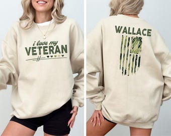 Puede incluir: Una sudadera beige con el texto "i love my VETERAN" en la parte delantera y el texto "WALLACE" en la parte posterior. La parte posterior también presenta un diseño de bandera estadounidense de camuflaje.