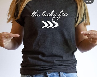 May include: Dark heather grey t-shirt with the text "the lucky few" in white script above three white chevron arrows. The model is wearing blue jeans and pointing at the shirt.