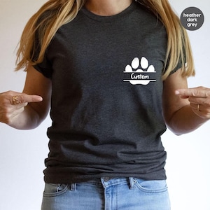 May include: Dark heather grey t-shirt with a white paw print design and the word "Custom" in a horizontal banner. The shirt is short-sleeved and made of a soft material. The shirt is paired with blue jeans.