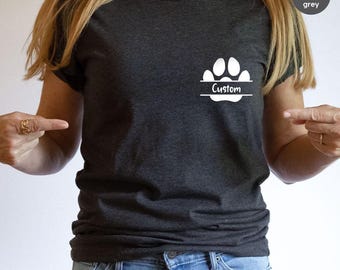 May include: Dark heather grey t-shirt with a white paw print design and the word "Custom" in a horizontal banner. The shirt is short-sleeved and made of a soft material. The shirt is paired with blue jeans.
