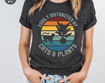 Puede incluir: Camiseta gris oscuro con un diseño gráfico circular. El diseño presenta una silueta de gato, una planta en maceta y el texto "EASILY DISTRACTED BY CATS & PLANTS" en un esquema de colores retro.