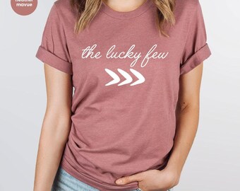 May include: A dusty rose t-shirt with rolled-up sleeves. The shirt features the text "the lucky few" in a cursive font, with three white arrow-like shapes below. The shirt is made of a soft material.