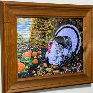 Può includere: Dipinto incorniciato di un tacchino in un campo di zucche e fogliame colorato. Il tacchino ha piume bianche, nere e blu, con un bargiglio rosso. L'opera d'arte è in una cornice di legno marrone, adatta per l'arredamento autunnale.