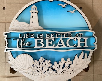 May include: A circular wooden wall sign featuring a layered design with a light blue sky, a lighthouse on a rocky shore, and two seagulls. A horizontal banner across the center displays the text LIFE IS BETTER AT the BEACH in white.