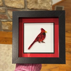以下が含まれることがあります： 赤いカージナルバードが描かれた額入りのアートワーク。鳥は細部までこだわって作られており、テクスチャ感があります。白い背景に赤い縁取りがあり、黒いフレームに入っています。インテリアの装飾品です。