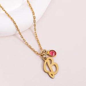 Op de afbeelding: Goudkleurige ketting met een hanger met de letter 'B' en een kleine, ronde, rode edelsteen. De fijne ketting ondersteunt de hanger, die een gestileerde letter is. De ketting wordt getoond op een wit oppervlak.