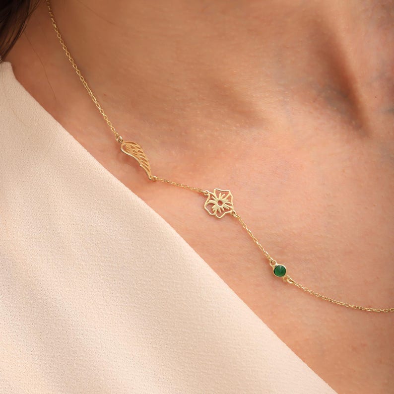 May include: A gold necklace with a wing charm, a flower pendant, and a small green gemstone. The necklace is shown on a person's neck, highlighting its elegant design and intricate details. The necklace is a beautiful piece of jewellery.