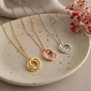 Op de afbeelding: Drie gepersonaliseerde kettingen met in elkaar grijpende ringen in goud, roségoud en zilver. Elke ring is gegraveerd met namen en data. De kettingen worden gepresenteerd op een getextureerd, gebroken wit oppervlak.