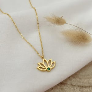 Op de afbeelding: Een goudkleurige ketting met een lotusbloemhanger. De hanger heeft een groene edelsteen in het midden. De ketting wordt gepresenteerd op een witte doek, met gedroogde planten op de achtergrond.