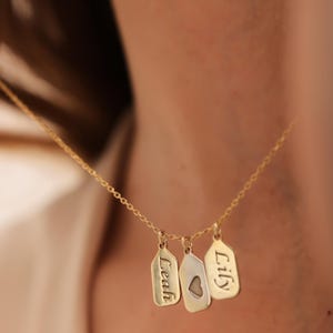 Op de afbeelding: Een goudkleurige ketting met drie hangertjes in de vorm van labels. De labels zijn gegraveerd met de namen "Leah" en "Lily", en één heeft een hartvormige uitsparing. De ketting is een fijne ketting, perfect voor dagelijks gebruik.