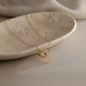 Op de afbeelding: Een delicate gouden ketting met twee in elkaar grijpende ringen. Eén ring is een volle cirkel, de andere heeft gegraveerde tekst. De ketting ligt op een lichtgekleurd, getextureerd oppervlak, wat een minimalistische en elegante uitstraling creëert.