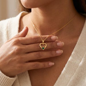 Op de afbeelding: Een goudkleurige ketting met een hartvormige hanger. De hanger heeft een hartvormige uitsparing en een kleine, donkergroene edelsteen. De ketting heeft een fijne ketting.