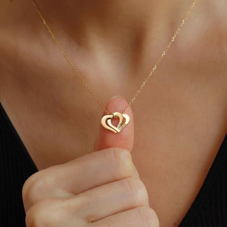 Op de afbeelding: Een goudkleurige ketting met een hanger bestaande uit twee in elkaar grijpende hartvormen. De hanger hangt aan een fijne goudkleurige ketting. Het oppervlak van de hartvormige hanger bevat een kleine gegraveerde tekst die zichtbaar is.