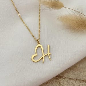 Op de afbeelding: Een goudkleurige ketting met een hanger in de vorm van een hart en de letter 'H'. De ketting is fijn en de hanger is uit één stuk. De ketting ligt op een witte stof.