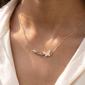 Op de afbeelding: Een gouden ketting met een verfijnde vlinderhanger ingelegd met kleine heldere steentjes, vergezeld door drie kleine ronde edelstenen in de kleuren lichtpaars, olijfgroen en roze, gerangschikt in een rij naast de vlinder.