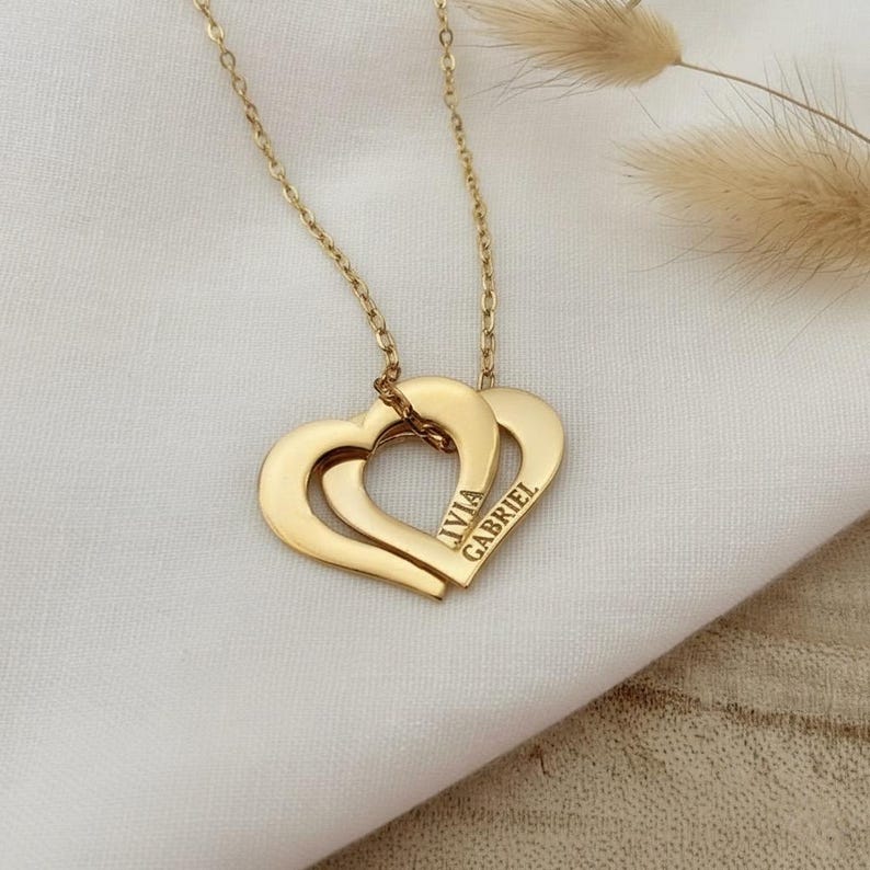 Op de afbeelding: Een goudkleurige metalen ketting met twee in elkaar grijpende hartvormige hangers. De hangers zijn gegraveerd met de namen IVIA en GABRIEL in een strak, schreefloos lettertype. De ketting hangt aan een fijne goudkleurige schakelketting.