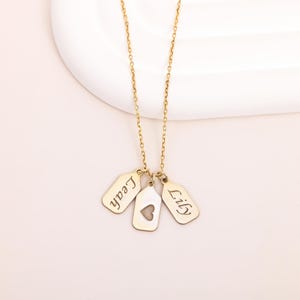 Op de afbeelding: Een goudkleurige ketting met drie hangertjes. De hangertjes zijn gegraveerd met de namen "Leah" en "Lily", en het middelste hangertje heeft een hartvormige uitsparing. De ketting wordt getoond op een witte ondergrond.