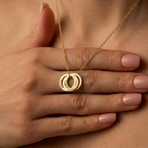 Op de afbeelding: Een goudkleurige ketting met drie in elkaar grijpende ringen. Elke ring heeft gegraveerde tekst. De ketting heeft een fijne ketting. De achtergrond is de huid van een persoon.