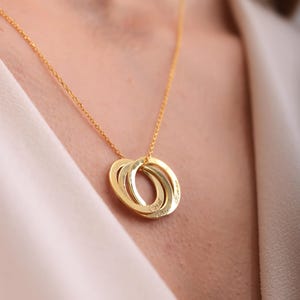 Può includere: Una collana dorata con tre anelli intrecciati, ognuno inciso con un nome. La collana è appesa a una catena sottile. Lo sfondo è di un colore morbido e neutro.